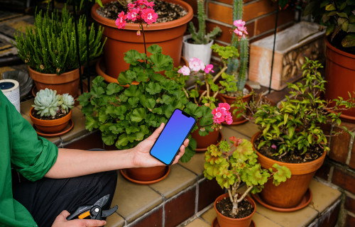 Mano femenina sosteniendo un iPhone 13 mockup en lugar ajardinado