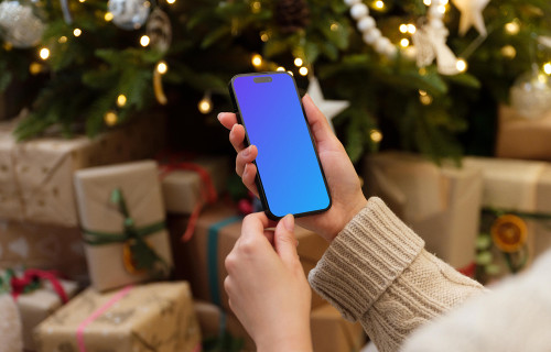 Mano sosteniendo un teléfono mockup junto al árbol de Navidad con regalos