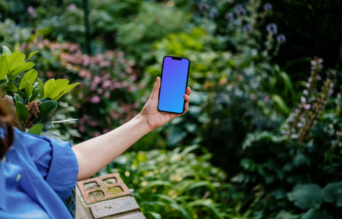 Mão segurando um iPhone 13 mockup cercado por plantas