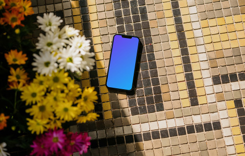iPhone 13 mockup en el pavimento de un jardín junto a las flores