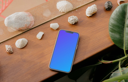 iPhone 13 Pro mockup sobre una mesa de madera junto a conchas marinas