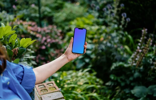 Hand holding an iPhone 13 mockup surrounded by plants