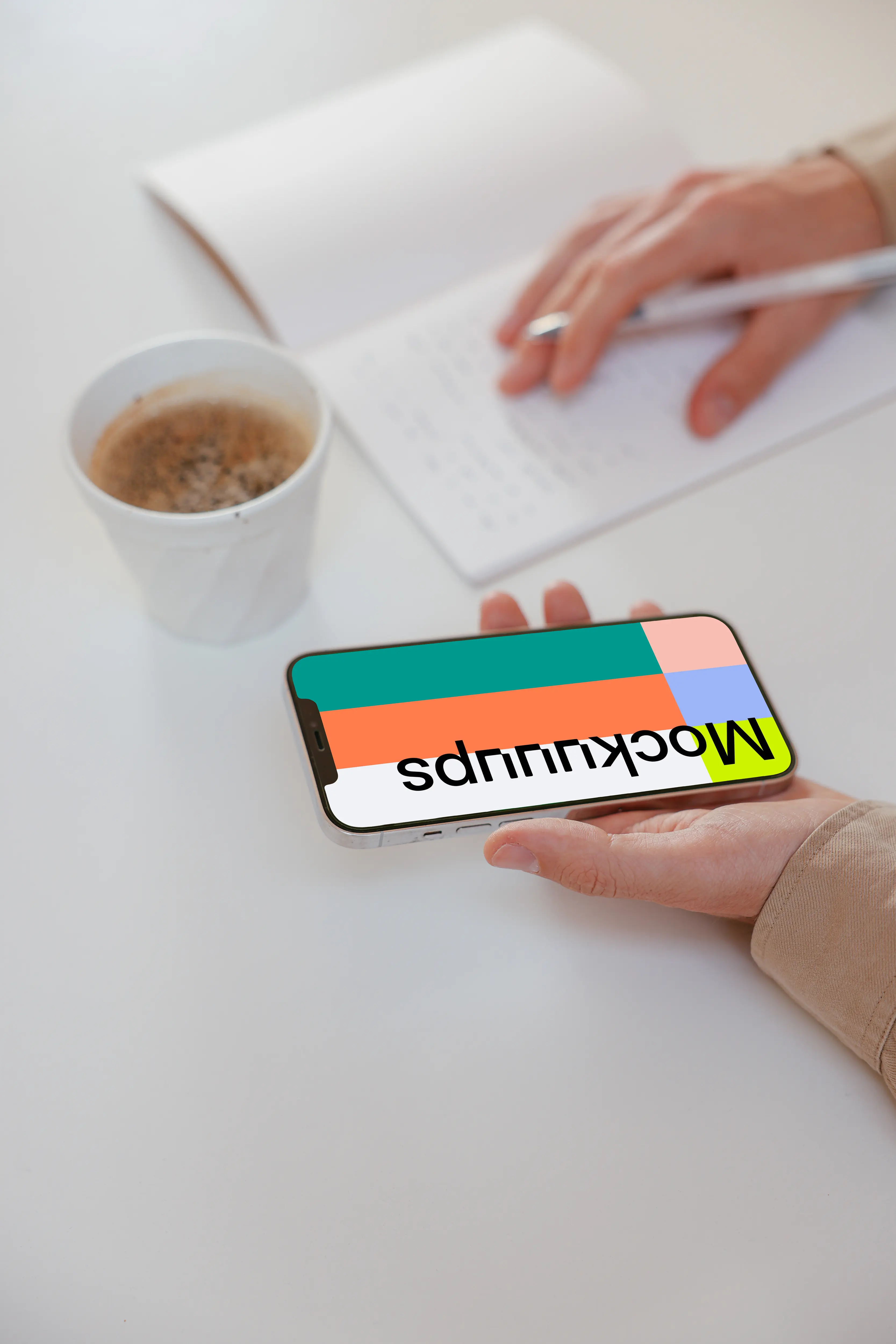 User holding iPhone and writing on a notebook beside a cup of coffee