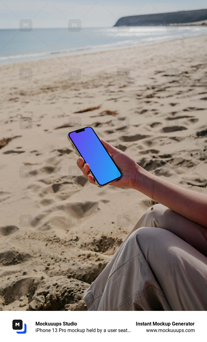 iPhone 13 Pro mockup held by a user seated on sand at the beach