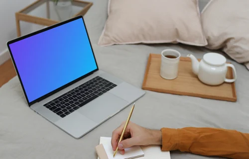 MacBook mockup on a bed with a user writing on a notebook