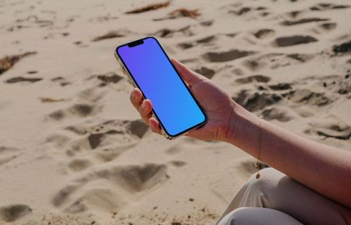 iPhone 13 Pro mockup sostenido por un usuario sentado en la arena de la playa