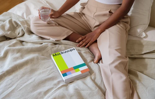 Book mockup on a cozy bed with a person holding a glass of water