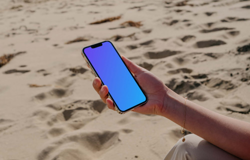 iPhone 13 Pro mockup held by a user seated on sand at the beach
