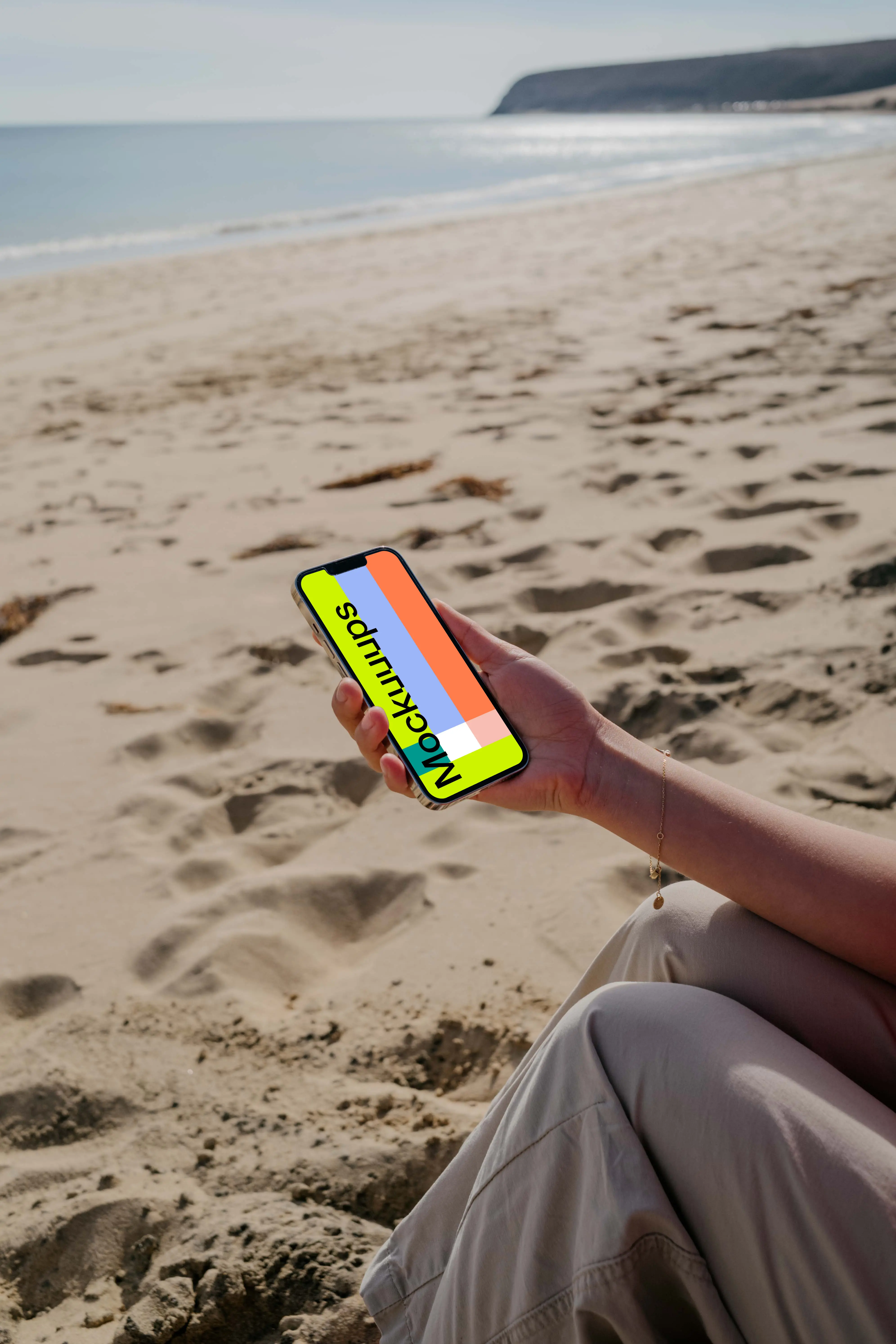 iPhone 13 Pro mockup held by a user seated on sand at the beach