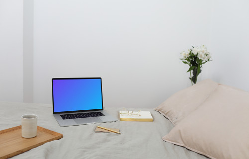 MacBook mockup on a bed with a book and wooden tray at the side.