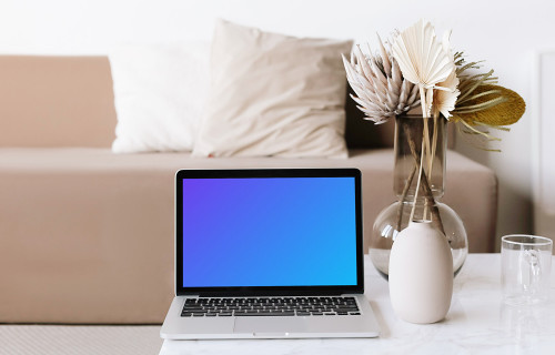 MacBook mockup on a white center table with glass cup and transparent flower po