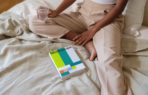 Book mockup on a cozy bed with a person holding a glass of water