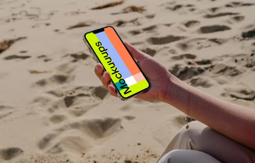 iPhone 13 Pro mockup held by a user seated on sand at the beach