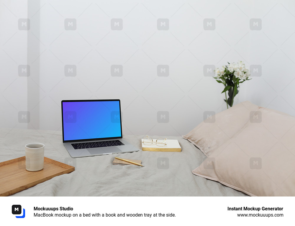 MacBook mockup on a bed with a book and wooden tray at the side.