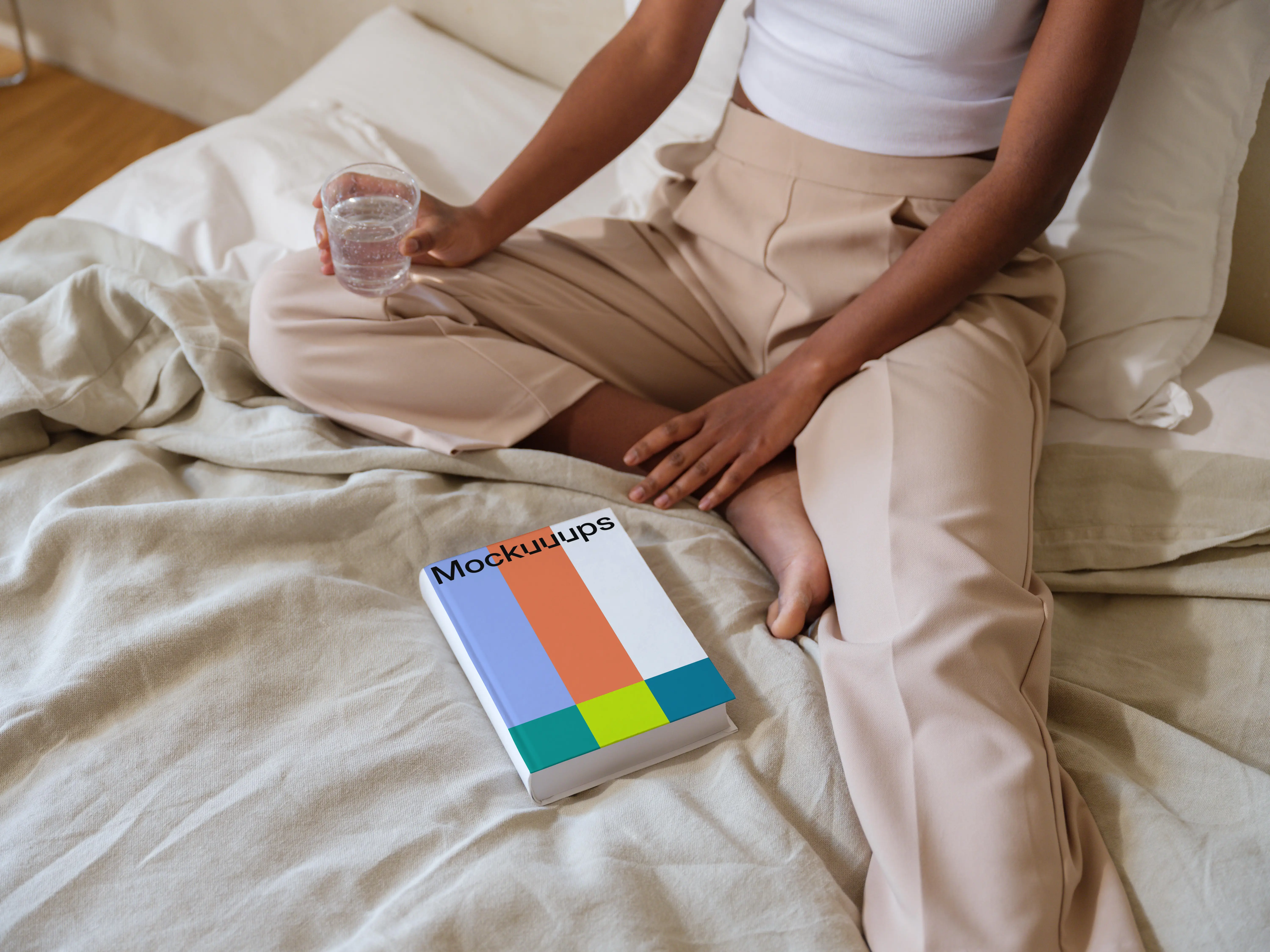 Book mockup on a cozy bed with a person holding a glass of water