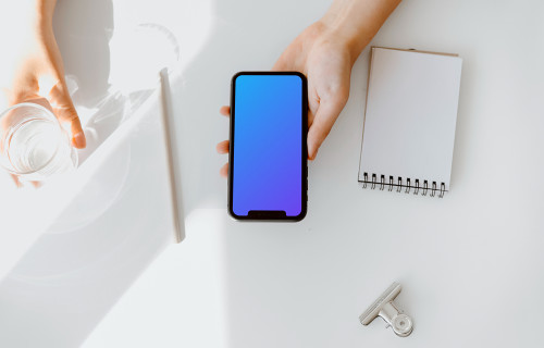 iPhone mockup held above a white table by a user with a glass of water at the side