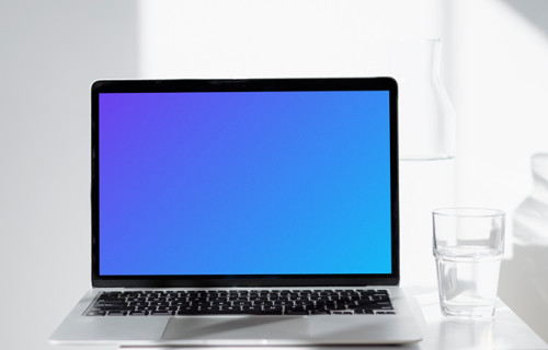 MacBook mockup sur une table blanche avec un verre d'eau à côté