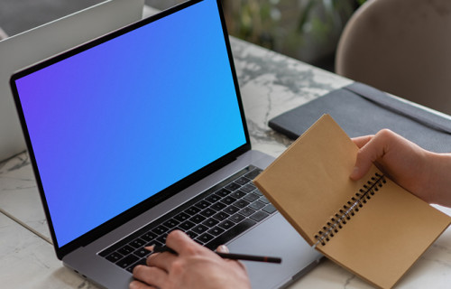 MacBook Pro mockup on a table with a jotter held beside it