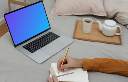 MacBook mockup on a bed with a user writing on a notebook