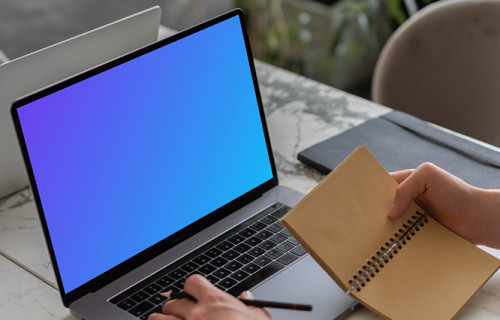 MacBook Pro mockup on a table with a jotter held beside it