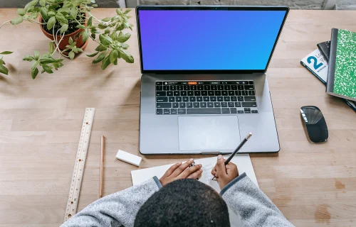 Young boy writing in a book with a MacBook Pro on the same table