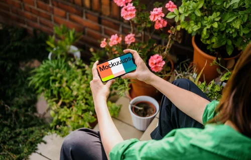 Woman sitting in garden while holding an iPhone 13 mockup