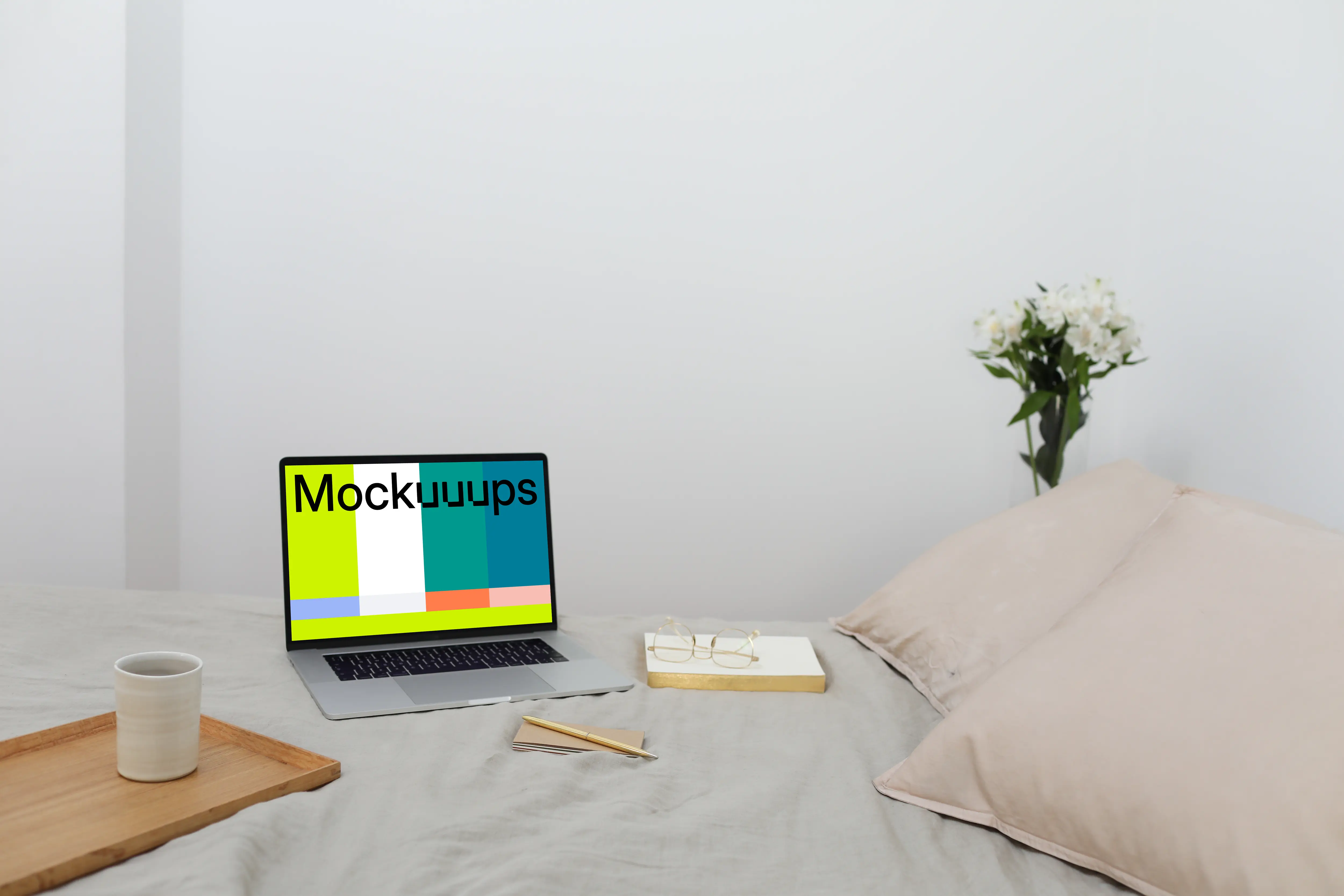 MacBook mockup on a bed with a book and wooden tray at the side.