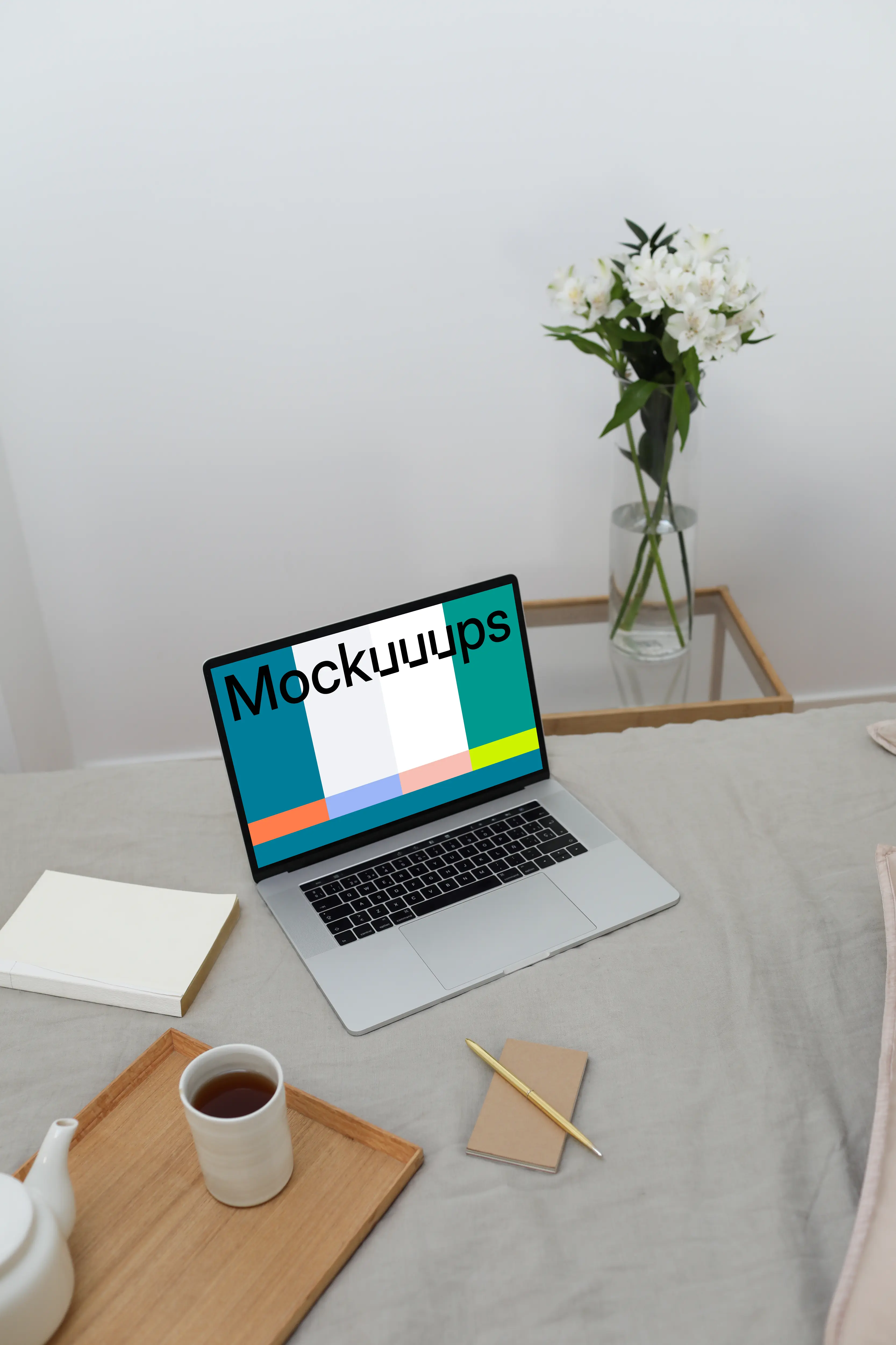 MacBook mockup on a bed with a jug and ceramic on a wooden tray and a notebook at the side