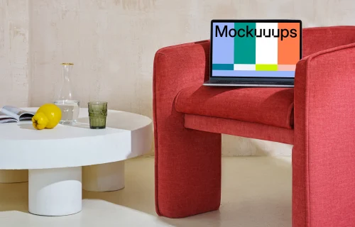 MacBook mockup at the edge of a red chair with a center table at the side