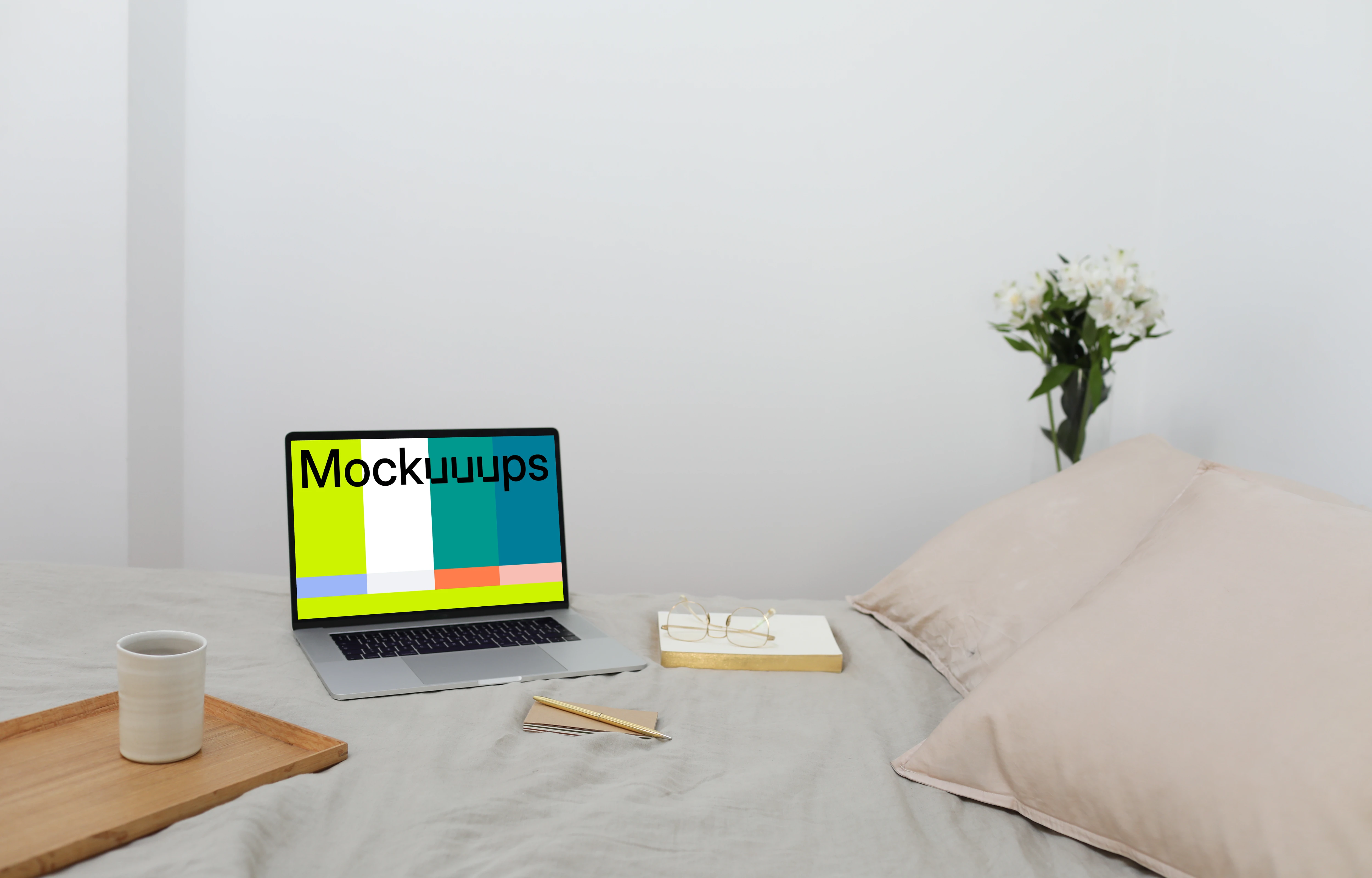 MacBook mockup on a bed with a book and wooden tray at the side.