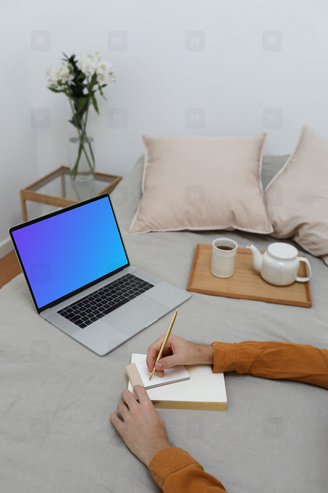 MacBook mockup on a bed with a user writing on a notebook